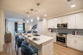 Located at 5050 Aspire Way in Naples, Florida, this thoughtfully designed one-bedroom, one-bathroom residence offers a well-appointed single-floor layout with modern finishes throughout. Light wood-style flooring flows seamlessly through the main living areas, complementing the bright, neutral color palette that defines the space.

The open living room is warm and inviting, featuring a ceiling fan, recessed lighting, and access to a private balcony that brings in natural light and outdoor views. The kitchen is a standout, outfitted with crisp white cabinetry accented by gold hardware, a blue-gray subway tile backsplash, quartz countertops, and a full suite of stainless steel appliances — including a refrigerator with water dispenser, electric range, and built-in microwave.

The bedroom is a comfortable retreat with plush carpeting, a ceiling fan, a window with outdoor views, and direct access to the bathroom. The bathroom is clean and modern, featuring a wide white vanity with gold hardware, a large mirror with overhead lighting, and light wood flooring that continues from the hallway. A dedicated closet and utility room provide practical storage solutions. The overall ambiance is contemporary and coastal, making it an ideal residence in the heart of Naples.