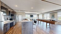 Located at 704 First Dr NW in Austin, Minnesota, this generously sized clubroom offers a well-appointed communal space with a modern industrial aesthetic. Warm hardwood-style flooring runs throughout, complemented by exposed metallic ductwork along the ceilings that adds an urban, loft-inspired character. Abundant natural light pours in through large windows fitted with roller shades, framing panoramic views of the surrounding cityscape and greenery. A door leads directly to an outdoor balcony, furnished with seating to enjoy the elevated vistas.

The space is organized into several distinct activity zones. A full kitchenette is anchored by a bold stone-accented feature wall, stainless steel appliances, dark cabinetry, and a granite-topped island with bar seating. A lounge area provides a comfortable sectional sofa and armchairs arranged around a striking tiled fireplace wall, above which a flat-screen TV is mounted. A dedicated game area accommodates both a shuffleboard table and a ping-pong table, each positioned near windows with wall-mounted TVs. A collaborative seating nook with a round table and modern chairs rounds out the amenities, making this clubroom a versatile and inviting gathering destination.