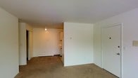 This single-floor unit presents a practical, open-concept layout that connects the main living area to the kitchen. The living space is finished with wall-to-wall carpeting in a neutral tone, lending a modest warmth to the interior. Clean white walls throughout keep the space feeling bright and versatile, providing a neutral backdrop adaptable to various furnishing styles.

A partial dividing wall separates the living area from the kitchen alcove, which features white cabinetry and is illuminated by a ceiling-mounted spotlight. A baseboard heater runs along the kitchen wall, indicating a straightforward heating system. The kitchen area transitions from carpet to a hard flooring surface, offering a practical distinction between the two zones.

A door on the right side of the main living area leads to a closet or additional storage space, contributing to the unit's overall functionality. The layout is efficiently organized, making good use of the available floor space. Overall, the unit offers a clean, no-frills environment well-suited for those seeking a simple and manageable living space.