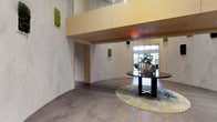 This sophisticated lobby space makes a striking first impression with its thoughtfully curated design elements and refined aesthetic. The most distinctive architectural feature is the sweeping curved wall clad in large-format marble-look panels, creating a fluid, organic flow throughout the entry area. Warm wood ceiling accents contrast beautifully against the lighter wall surfaces, adding depth and a sense of understated luxury to the space.

Natural light floods in through floor-to-ceiling windows and glass entry doors at the far end, creating a bright and inviting atmosphere. Living moss wall panels are strategically placed as green accent features, introducing a touch of nature and vibrancy against the neutral palette. The flooring is a contemporary wide-plank hardwood in a cool gray tone, providing a clean, modern foundation that ties the design together.

At the center of the space, a round dark wood table sits atop a circular area rug in soft gold and blue tones, styled with decorative vases and a potted plant. This arrangement serves as a natural focal point, anchoring the room and adding warmth. The overall ambiance is polished and professional, balancing clean modern architecture with inviting, nature-inspired touches — ideal for a commercial or hospitality setting.