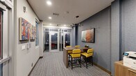 Located at 3200 Brighton Blvd in Denver, CO, this compact yet efficiently designed collaborative workspace offers a thoughtfully curated environment ideal for focused work and small group meetings. The single-floor layout maximizes every square foot, seamlessly blending multiple functional zones within a cohesive design.

The main area features a central high-top communal table paired with bold yellow upholstered counter chairs, anchored beneath a striking multi-arm sputnik chandelier. A textured blue-grey accent wall provides a sophisticated backdrop, adorned with a large-format framed artwork that adds a vibrant, urban aesthetic. A mounted flat-panel display enhances the space's utility for presentations or collaborative work.

Along one wall, a set of glass-paneled private phone booths offer semi-enclosed spaces for focused, individual use, each equipped with seating and interior lighting. A secondary seating nook with colorful patterned cushions and an additional work surface provides yet another flexible option for informal meetings or solo work.

Practical amenities include a shared printer station set on a warm wood-toned cabinet. Frosted glass windows and interior partitions maintain privacy while allowing light to circulate throughout. The space is finished with multi-toned loop carpet, recessed lighting, and curated wall art, creating a modern, professional, and energizing atmosphere throughout.