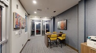 Located at 3200 Brighton Blvd in Denver, CO, this compact yet efficiently designed collaborative workspace offers a thoughtfully curated environment ideal for focused work and small group meetings. The single-floor layout maximizes every square foot, seamlessly blending multiple functional zones within a cohesive design.

The main area features a central high-top communal table paired with bold yellow upholstered counter chairs, anchored beneath a striking multi-arm sputnik chandelier. A textured blue-grey accent wall provides a sophisticated backdrop, adorned with a large-format framed artwork that adds a vibrant, urban aesthetic. A mounted flat-panel display enhances the space's utility for presentations or collaborative work.

Along one wall, a set of glass-paneled private phone booths offer semi-enclosed spaces for focused, individual use, each equipped with seating and interior lighting. A secondary seating nook with colorful patterned cushions and an additional work surface provides yet another flexible option for informal meetings or solo work.

Practical amenities include a shared printer station set on a warm wood-toned cabinet. Frosted glass windows and interior partitions maintain privacy while allowing light to circulate throughout. The space is finished with multi-toned loop carpet, recessed lighting, and curated wall art, creating a modern, professional, and energizing atmosphere throughout.