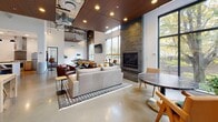 Located at 337 NE 103rd St in Seattle, WA, this single-floor communal amenity space offers a well-appointed environment designed for both relaxation and social interaction. The open-plan layout is anchored by soaring ceilings that amplify the sense of space, with exposed ductwork and industrial-style pendant lighting adding an urban-loft aesthetic. Polished concrete floors run throughout, complementing the warm wood slat ceiling panels that define the lounge area.

The main gathering space features a full-size pool table as a central focal point, surrounded by thoughtfully arranged seating that includes a plush sofa, leather armchairs, and a patterned area rug. A dramatic floor-to-ceiling stone fireplace provides a cozy anchor to the lounge, flanked by large floor-to-ceiling windows that invite abundant natural light and offer views of the surrounding greenery. A wall-mounted TV and additional dining-height seating with bar stools round out the entertainment options.

The kitchen is finished to a high standard, featuring light wood cabinetry, white quartz countertops, subway tile backsplash, stainless steel appliances including a range, dishwasher, and refrigerator, as well as open shelving for added functionality. This space delivers a refined yet inviting atmosphere ideal for community gatherings.