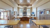 This single-floor space is a well-appointed demonstration kitchen and multipurpose gathering area designed for both culinary instruction and social entertaining. The kitchen is anchored by a large butcher block central island and equipped with a professional stainless steel range, an oversized stainless steel ventilation hood, and ample cabinetry with light wood finishes and white countertops. Multiple windows with black-framed grid panes line the walls, inviting generous natural light and views of surrounding greenery. A wall-mounted display screen provides a functional focal point for demonstrations or presentations.

The main gathering area features soaring ceilings with a distinctive stepped, wood-paneled ceiling detail and recessed and pendant lighting. Warm hardwood floors run throughout, complementing the light, neutral tile wall cladding. Tall communal tables paired with industrial-style bar stools offer flexible seating for participants or guests. Decorative wine barrel medallions add character to the walls.

Large sliding barn-style doors connect the kitchen and gathering area to an adjacent lounge, which houses a billiards table and comfortable seating, creating a seamless flow between spaces. The overall ambiance is warm, modern, and purposefully designed for both hands-on culinary activity and relaxed social interaction.