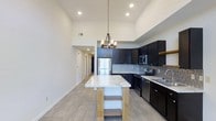 This single-floor residence showcases a thoughtfully designed open-concept layout that blends modern aesthetics with functional living. The kitchen serves as the centerpiece of the space, featuring striking dark cabinetry that extends nearly to the ceiling, creating a bold contrast against the bright white walls. A freestanding kitchen island with a veined marble countertop and open lower shelving provides both prep space and a casual gathering spot, while a rustic wood-and-metal chandelier overhead adds a warm, inviting touch. Stainless steel appliances, including a refrigerator, microwave, and range, lend a sleek, contemporary finish, complemented by a decorative tile backsplash that adds texture and visual interest. The sink area features additional marble countertops, maintaining a cohesive design throughout. Light wood-toned flooring runs throughout the space, enhancing the sense of openness and warmth. Soaring ceilings amplify the airy feel of the home, while recessed lighting provides ample illumination. A hallway visible from the kitchen connects to additional areas of the residence, suggesting a well-organized and efficiently planned floor plan throughout.
