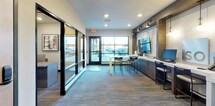 This single-floor commercial space is designed as a polished leasing or property management office, combining functionality with a contemporary aesthetic. The layout flows openly from a welcoming entryway corridor into the main workspace, creating a clear sense of arrival and direction for visitors.

The entry hallway is lined with an eye-catching, large-format neighborhood map mural that serves as a visual focal point, complemented by framed artwork and a small coffee station on a white console, adding a hospitality-forward touch. A pair of teal-framed glass double doors connects the corridor to the exterior, allowing natural light to filter through.

The main office area is anchored by a central communal table with seating, positioned beneath recessed lighting. A built-in dark wood workstation runs along one wall, housing monitors and business equipment, with pendant lighting adding warmth. Large storefront-style windows along the front facade provide ample natural light and views of the surrounding parking area and exterior.

A glass-partitioned private office sits adjacent to the main floor, maintaining visual openness while providing a dedicated, enclosed workspace. Durable patterned carpet tiles in neutral tones ground the entire space, contributing to a clean, professional ambiance throughout.