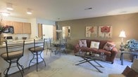 This single-floor residence offers an open-concept layout that connects the living, dining, and kitchen areas in a cohesive and functional design. The living area is anchored by a plush sofa set against a warm taupe accent wall, complemented by a glass-top coffee table and framed botanical artwork that adds a decorative touch. The space is finished with soft wall-to-wall carpeting, creating a comfortable and inviting atmosphere. Adjacent to the living area, the dining space features a round table with sleek metal chairs, illuminated by a pendant light fixture that draws the eye upward and adds a modern accent. A breakfast bar with bar stools creates a casual dining alternative and serves as a natural divider between the dining area and the kitchen. The kitchen is visible in the background, offering a functional and accessible layout. Neutral tones throughout the space — from the warm beige walls to the light-toned flooring in the kitchen — create a unified aesthetic that feels both clean and welcoming. Ceiling-mounted HVAC vents and recessed lighting ensure year-round comfort, while the open sightlines between rooms enhance the overall sense of space and flow.