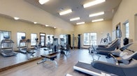 This fitness center is a thoughtfully designed workout facility that offers a comprehensive range of exercise equipment within a bright, open environment. The space features hardwood-style flooring throughout, providing a clean and durable surface suited for high-traffic use. Large windows along the walls allow abundant natural light to fill the room, creating an energizing atmosphere for workouts.

The facility is outfitted with an extensive selection of strength training machines, including cable systems, weight benches, and multi-function resistance units, catering to a full range of muscle groups. Cardio equipment such as ellipticals, stationary bikes, and treadmills are also prominently featured, ensuring a well-rounded fitness experience.

Full-length mirrored walls line one side of the room, enhancing the sense of space while allowing users to monitor their form during exercise. Overhead fluorescent lighting supplements the natural light, ensuring the space remains well-lit at all times. The elevated ceiling height contributes to an open, airy feel that prevents the room from feeling cramped even when fully equipped. Overall, this fitness center delivers a functional, organized, and inviting environment designed to support a wide variety of workout routines.