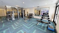 The fitness center is a thoughtfully designed workout space that combines functionality with a modern aesthetic. The room features a striking herringbone-patterned carpet in teal and grey tones, providing both visual interest and a durable, cushioned surface underfoot. Recessed lighting in the white ceiling ensures the space is well-illuminated throughout.

Strength training is well-represented, with a Matrix cable machine offering versatile resistance exercise options. A multi-tiered dumbbell rack holds a comprehensive range of free weights, accompanied by adjustable weight benches nearby for a variety of pressing and curling exercises. A medicine ball rack stocked with multiple weighted balls supports functional and core training routines.

A large wall-mounted mirror expands the sense of space while allowing users to monitor their form during workouts. The mirror reflects an additional adjoining area, suggesting the fitness facility extends further, offering even more room for exercise. Neutral-toned walls and clean, white-painted finishes contribute to a bright and motivating atmosphere, making this fitness center a practical and inviting space for residents or guests to pursue their wellness goals.
