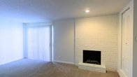 This single-floor space presents a clean and understated interior centered around a striking white-painted brick fireplace, which serves as the focal point of the main living area. The fireplace features a classic hearth with a black firebox and a grate, extending floor-to-ceiling along the wall and adding a sense of architectural character. The living area is finished with wall-to-wall neutral carpeting, contributing to a warm and comfortable atmosphere. A large sliding glass door fitted with vertical blinds allows natural light to filter into the space while providing potential access to an outdoor area. The walls are painted in a crisp white, complementing the bright, airy feel of the interior. Recessed lighting in the ceiling provides supplemental illumination. The textured ceiling adds a subtle decorative element consistent with the overall design. The layout feels open and versatile, lending itself well to a variety of furniture arrangements. The combination of the brick fireplace, soft carpeting, and ample natural light creates a balanced and inviting environment suited for comfortable everyday living.