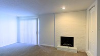 This single-floor space presents a clean and understated interior centered around a striking white-painted brick fireplace, which serves as the focal point of the main living area. The fireplace features a classic hearth with a black firebox and a grate, extending floor-to-ceiling along the wall and adding a sense of architectural character. The living area is finished with wall-to-wall neutral carpeting, contributing to a warm and comfortable atmosphere. A large sliding glass door fitted with vertical blinds allows natural light to filter into the space while providing potential access to an outdoor area. The walls are painted in a crisp white, complementing the bright, airy feel of the interior. Recessed lighting in the ceiling provides supplemental illumination. The textured ceiling adds a subtle decorative element consistent with the overall design. The layout feels open and versatile, lending itself well to a variety of furniture arrangements. The combination of the brick fireplace, soft carpeting, and ample natural light creates a balanced and inviting environment suited for comfortable everyday living.
