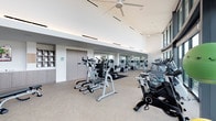 This spacious fitness center is designed with a clean, contemporary aesthetic that prioritizes both functionality and comfort. The facility is thoughtfully organized to accommodate a wide range of workout routines, offering an extensive array of equipment including treadmills, elliptical trainers, stationary and spin bikes, cable machines, free weights, and strength-training stations.

Architecturally, the space benefits from soaring ceilings fitted with large ceiling fans and recessed linear lighting that ensures consistent, even illumination throughout. Floor-to-ceiling glass windows line one side of the facility, flooding the interior with abundant natural light and providing expansive views of the surrounding outdoor terrace, greenery, and the city skyline beyond — an inspiring backdrop for any workout.

The interior finishes are refined and understated, featuring neutral-toned carpeted flooring, crisp white walls, and built-in cabinetry with open shelving along one wall, adding practical storage to the space. A full-length mirrored wall expands the visual depth of the main workout area. The overall ambiance is bright, airy, and professional, reflecting a high-quality facility designed to support an active lifestyle.