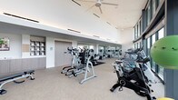 This spacious fitness center is designed with a clean, contemporary aesthetic that prioritizes both functionality and comfort. The facility is thoughtfully organized to accommodate a wide range of workout routines, offering an extensive array of equipment including treadmills, elliptical trainers, stationary and spin bikes, cable machines, free weights, and strength-training stations.

Architecturally, the space benefits from soaring ceilings fitted with large ceiling fans and recessed linear lighting that ensures consistent, even illumination throughout. Floor-to-ceiling glass windows line one side of the facility, flooding the interior with abundant natural light and providing expansive views of the surrounding outdoor terrace, greenery, and the city skyline beyond — an inspiring backdrop for any workout.

The interior finishes are refined and understated, featuring neutral-toned carpeted flooring, crisp white walls, and built-in cabinetry with open shelving along one wall, adding practical storage to the space. A full-length mirrored wall expands the visual depth of the main workout area. The overall ambiance is bright, airy, and professional, reflecting a high-quality facility designed to support an active lifestyle.