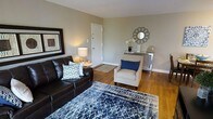 This single-floor living space showcases a thoughtfully arranged living room that balances comfort and style. The room features warm hardwood flooring that extends throughout, complemented by neutral greige walls that create a calm, welcoming atmosphere. Natural light enhances the space, highlighting the carefully chosen furnishings and décor.

The living area is anchored by a dark leather sofa adorned with patterned throw pillows in navy and white, paired with a neutral upholstered accent chair that adds a classic touch. A bold navy and white geometric area rug grounds the seating arrangement, adding depth and visual interest to the room.

A mirrored console table along one wall serves as an elegant focal point, styled with decorative accents including a sunburst mirror, floral arrangement, and curated accessories. A framed art piece on the adjacent wall adds a sophisticated finishing touch. A white interior door provides access to additional areas of the space, while a patterned entryway rug near the entrance adds a practical yet decorative element. Overall, the space presents a cohesive, polished aesthetic with attention to detail throughout.