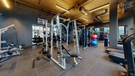 Located at 633 South LaSalle Street in Chicago's South Loop, this fitness facility offers a comprehensive, well-appointed workout environment spread across a single open floor. The space features an industrial-chic aesthetic, with exposed ductwork and piping running along the ceiling, complemented by sleek linear LED lighting that illuminates the facility throughout.

Large floor-to-ceiling windows line the perimeter, flooding the space with natural light and offering views of the surrounding Chicago skyline. The flooring is covered with durable rubber gym tiles throughout, providing both safety and functionality.

The facility is thoroughly equipped for strength training, featuring multiple power racks and squat stands loaded with Olympic barbells and weight plates, a Smith machine, a full rack of dumbbells ranging across various weights, and adjustable benches. A dedicated functional training area includes kettlebells, battle ropes, a punching bag, and TRX suspension trainers. Stability balls and additional accessories are also available for varied workouts.

Full-length mirrors are strategically positioned along the walls to assist with form and to enhance the sense of space. Cardio equipment, including a treadmill and a stationary bike, rounds out the offerings. The overall ambiance is modern, focused, and purpose-built to support a wide range of fitness goals.