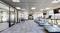 Located at 955 W Clairemont Ave in Eau Claire, Wisconsin, this well-appointed fitness center offers a spacious, single-floor layout designed to support a comprehensive workout experience. The facility is housed in a bright, open room finished with a contemporary dropped tile ceiling equipped with recessed panel lighting that ensures consistent, even illumination throughout the space.

The flooring consists of patterned, multi-tone carpet tiles that add a modern aesthetic while providing comfort underfoot. Large windows line multiple walls, inviting generous amounts of natural light and offering views of the surrounding outdoor greenery, creating an open and energizing atmosphere.

The equipment is thoughtfully arranged to maximize usable floor space. A dedicated cardio zone features a row of treadmills positioned along the window-facing wall, alongside elliptical trainers, stationary bikes, and a rowing machine. A free weights area includes a tiered dumbbell rack with an accompanying flat bench and additional benching options, while a cable resistance machine supports functional strength training. A stability ball and additional accessories round out the strength training offerings.

Wall-mounted flat-screen televisions are positioned throughout the room for entertainment during workouts. A floor-to-ceiling glass partition wall with dark-framed doors creates a transparent division, connecting the fitness area to an adjacent space. The overall ambiance is clean, modern, and professional, making it a functional and inviting environment for fitness.
