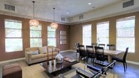 This single-floor space offers a thoughtfully designed open-concept layout that seamlessly blends a living area with a formal dining space. The warm, earthy tones of the walls create a welcoming and grounded ambiance throughout. The living area is anchored by a plush neutral sofa and a dark wood coffee table, accessorized with decorative accents including a striking glass vase, adding a refined yet approachable aesthetic. Two ornate chain-hung pendant light fixtures serve as a focal point, casting a warm, ambient glow over the seating area. The dining area is furnished with a dark wood table surrounded by matching high-back chairs, complemented by a candelabra centerpiece that adds a touch of elegance. Large windows fitted with natural woven shades line multiple walls, flooding the space with generous natural light while offering views of lush greenery outside. Crown molding runs along the ceiling perimeter, adding a classic architectural detail, while recessed lighting and tile flooring with a large area rug complete the polished, cohesive interior. The overall atmosphere is sophisticated yet comfortable, making the space well-suited for both everyday living and entertaining.