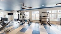This state-of-the-art fitness center, located at 3830 NC-55 E in Durham, NC, spans 1,160 square feet on a single floor. The space is designed to maximize functionality and aesthetic appeal. The room features a striking blue ceiling with modern lighting fixtures and ceiling fans, creating a cool, energizing atmosphere. Large windows line the walls, flooding the area with natural light and offering views of the surrounding property.

The flooring is a distinctive checkerboard pattern in blue and beige, adding visual interest and demarcating different workout zones. The center is equipped with a comprehensive range of exercise machines, including treadmills, ellipticals, stationary bikes, and weight training equipment. A dedicated area for free weights and resistance training is visible, complete with mirrors for form checking.

The layout is thoughtfully arranged to accommodate various workout styles and equipment types. Wall-mounted TVs provide entertainment options, while water fountains and towel stations offer convenience. The overall design balances practicality with a modern, inviting ambiance, making it an ideal space for fitness enthusiasts of all levels.