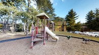 This outdoor playground space offers a welcoming recreational area surrounded by mature evergreen and deciduous trees that provide natural shade and a lush, park-like atmosphere. The centerpiece of the space is a classic multi-level play structure featuring a covered platform tower with a enclosed tube slide, climbing stairs, and colorful support poles in red and pink tones. The structure sits atop a wood chip safety surface, which cushions the play area and defines its boundaries. A small spring-rider toy is also visible nearby, adding an additional play element for younger children. The grounds surrounding the play area transition into open grassy lawn, creating additional space for outdoor activities. Tall conifers frame the setting, lending a sense of enclosure and tranquility. A multi-story residential building is visible in the background, situating this playground as an amenity within a larger residential community. The open sky above and natural tree canopy contribute to a bright, airy environment that feels both safe and inviting for children and families alike.