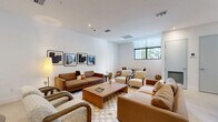 Located at 2555 NE 179th St in North Miami Beach, FL, this single-floor multipurpose amenity space offers a thoughtfully designed environment suited for both relaxation and collaboration. The open layout is anchored by a generously sized lounge area furnished with a mix of leather and upholstered sofas, accent armchairs, and a solid wood coffee table atop a checkered area rug, creating a warm, inviting atmosphere. A series of framed abstract artworks lines the crisp white walls, adding a curated, gallery-like quality to the space. Large black-framed windows allow natural light to filter in, offering views of the surrounding greenery and enhancing the airy feel of the interior. Recessed lighting and floor lamps provide layered illumination throughout. A defined meeting or conference zone features a sizable light wood table surrounded by upholstered chairs, ideal for structured gatherings or work sessions. A textured acoustic wall panel serves as a refined backdrop in this area. Neutral-toned large-format tile flooring flows throughout, tying together the lounge and meeting zones seamlessly. The clean, minimalist aesthetic — characterized by white walls, warm wood tones, and soft neutral furnishings — reflects a modern yet comfortable design sensibility well-suited to the North Miami Beach lifestyle.