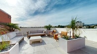 Located at 11600 Santa Monica Boulevard in the heart of West Los Angeles, this rooftop terrace offers a sophisticated and well-appointed outdoor amenity space with striking views of the surrounding cityscape and distant mountain ranges. The terrace is finished with large-format porcelain tile pavers throughout, lending a clean, contemporary aesthetic to the open-air environment.

The space is organized into several distinct zones designed for relaxation and social gathering. A primary lounge area features a cushioned sofa, teak-framed armchairs, and a low-profile coffee table, all arranged to take full advantage of the panoramic Los Angeles skyline views. A separate turf-covered seating area provides a softer, more casual setting, complete with teak chairs and an outdoor grill — ideal for entertaining.

Lush, thoughtfully placed landscaping defines the rooftop's character. Oversized white planters are filled with tropical palms, agaves, ornamental grasses, and small trees, creating natural dividers between zones while adding vibrant greenery against the open sky. A water feature and decorative stone accents add refined detail throughout.

Warm terracotta-toned architectural panels and slatted wood privacy screens provide visual interest and partial enclosure, while the open perimeter showcases unobstructed views of the West Los Angeles urban landscape. This rooftop terrace delivers a compelling blend of modern design, curated landscaping, and exceptional outdoor livability.