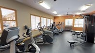 This fitness center is a comprehensively outfitted single-level space designed to support a full range of workout routines. The interior is finished with warm beige and bold orange accent walls trimmed with wood detailing, creating an energetic yet welcoming atmosphere. Dark rubber flooring spans the entire space, providing a durable, gym-appropriate surface throughout.

The cardio section is well-stocked with multiple elliptical trainers, stationary bikes, and treadmills from brands including Matrix and True Fitness, arranged in organized rows along the windowed wall. Multiple wood-framed windows fitted with horizontal blinds line the perimeter, allowing natural light to filter in while maintaining privacy.

A large wall-mounted mirror runs the length of one side of the room, visually expanding the space and allowing users to monitor their form during workouts. The strength training area includes a cable machine, weight bench, and a tiered dumbbell rack holding a full range of free weights. A wall-mounted flat-screen television is positioned for easy viewing from multiple workout stations.

Overhead, flush-mounted fluorescent lighting ensures consistent illumination, while ceiling fans promote air circulation throughout the space. The fitness center also features glass-paneled entry doors, adding a bright, open feel to the entrance area.