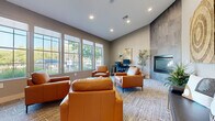 Located at 9001 Renner Blvd in Lenexa, Kansas, this single-floor leasing center for Estancia offers a thoughtfully designed space that balances professional function with residential warmth. Visitors are greeted at the entrance by light hardwood flooring, glass-paneled doors, and branded signage, setting a polished first impression. The main lounge area transitions seamlessly from the entry, featuring plush cognac leather armchairs arranged around a rustic round coffee table, all set atop a layered area rug. A standout feature of the space is the floor-to-ceiling large-format tile fireplace surround, which anchors the room with a sleek, contemporary aesthetic. Adjacent to the fireplace, a dedicated workstation with a desktop computer provides a functional area for leasing consultations. A bank of large, grid-paned windows lines the exterior wall, flooding the interior with natural light and offering clear views of the surrounding greenery. Recessed lighting throughout the ceiling complements the natural light, ensuring a bright and inviting atmosphere at all times. Decorative wall art, including two circular woven panels, adds an organic, textural element to the neutral-toned walls. The overall ambiance is modern, comfortable, and professional, making this leasing center a welcoming destination for prospective residents.