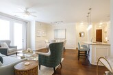 This single-floor residence combines refined architectural details with a warm, cohesive aesthetic throughout. The entryway welcomes with rich hardwood flooring, crisp white walls, and crown molding that carry through the main living areas. The living room is bright and inviting, benefiting from large windows with floor-to-ceiling drapes and a ceiling fan, creating an airy atmosphere ideal for relaxing or entertaining. Adjacent to the living room, a dedicated dining area features elegant wainscoting on the walls and track lighting overhead, lending a polished, formal character to the space. The kitchen is well-equipped with white shaker-style cabinetry, granite countertops, a classic subway tile backsplash, and a full suite of stainless steel appliances, including a refrigerator, range, and microwave. A central island provides additional prep and serving space. The bedroom is generously sized, carpeted for comfort, and offers ample natural light through a large window. A walk-in closet provides substantial storage, and a wall-mounted television is included. The bathroom is finished with a dual-sink granite vanity, white cabinetry, and a glass-enclosed shower, maintaining the home's clean and sophisticated style throughout.