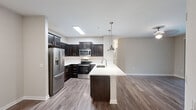 This single-floor residence is designed with a contemporary open-concept layout that seamlessly connects the kitchen and living areas. The kitchen is a standout feature, equipped with rich dark-espresso cabinetry, a white quartz peninsula with an undermount stainless steel sink, and stainless steel appliances including a French door refrigerator, electric range, built-in microwave, and dishwasher. A white subway tile backsplash adds a clean, modern contrast, while two brushed nickel pendant lights illuminate the peninsula. Wood-look plank flooring runs throughout the main living area, enhancing the open feel of the space. A sliding glass door leads to a private balcony, allowing natural light to fill the living area. The bedroom features plush carpet, a ceiling fan, and dual windows fitted with horizontal blinds for privacy and light control. A walk-in closet with wire shelving provides generous storage. The bathroom includes tile flooring, a glass-enclosed shower with tile surround, a white quartz vanity top with dark cabinetry, and a well-lit mirror. A dedicated laundry area accommodates a full-sized washer and dryer.