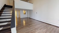 Located at Silverbrick Lofts in Springfield, MA, this two-story loft unit combines modern finishes with an open, airy design. The main living area immediately impresses with its dramatic double-height ceilings and a mezzanine level overlooking the space below, creating a sense of verticality and openness. Warm hardwood-style flooring runs throughout both levels, lending cohesion and a clean aesthetic to the entire unit. The spacious main floor living area is bright and open-plan, with baseboard heating and a pass-through window connecting to the kitchen. The kitchen is efficiently laid out in a galley style, featuring white cabinetry with contemporary hardware, dark countertops, a subway tile backsplash, and stainless steel appliances including a dishwasher and refrigerator. Two bathrooms serve the unit — one with a tiled tub-shower combination, dark wood vanity, and neutral tile flooring, and a second powder room with a matching vanity and mirror. The upper loft level, accessible via a staircase, offers an intimate space with a sloped ceiling and exposed wood beam detail, ideal for use as a bedroom or private retreat. A window with vertical blinds allows natural light to filter into the upper level. The overall design is clean, modern, and well-suited for everyday living.