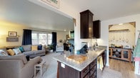 This single-story home offers a well-designed, open-concept layout that seamlessly connects the kitchen and living spaces. The kitchen is outfitted with dark espresso cabinetry, granite countertops, stainless steel appliances, and a central island with a sink — ideal for both cooking and casual entertaining. Light hardwood-style flooring runs through the kitchen, while a separate dining nook adds additional functionality. A dedicated butler's pantry or serving area with pendant lighting is accessible from the kitchen.

The living room is generously sized with plush carpeting, multiple windows dressed in navy drapes, and a sliding glass door that fills the space with natural light. Two bedrooms feature ceiling fans, carpeted floors, and windows with outdoor views. The primary bedroom includes mirrored closet doors, while the secondary bedroom offers a bright window framed by patterned curtains.

Two full bathrooms are finished with granite-topped dark vanities and tiled surrounds — one with a glass-enclosed walk-in shower and another with a tub-shower combination. Outside, a white-railed deck overlooks a well-maintained yard with mature trees and open green space, adding a pleasant outdoor retreat to the home.