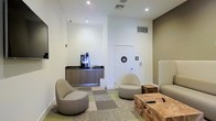 This single-floor commercial space offers a clean, modern design with a thoughtfully arranged lounge area that sets a professional yet comfortable tone. The lounge features contemporary pod-style seating chairs in neutral tones, paired with a rustic natural wood coffee table that adds an organic contrast to the otherwise sleek interior. A patterned carpet in muted greens and grays defines the seating area, providing a subtle visual anchor within the open layout.

A dedicated refreshment station lines one wall, outfitted with a dark countertop, warm wood cabinetry, and a professional-grade coffee machine, making it a convenient amenity for visitors and occupants alike. Recessed ceiling lighting illuminates the space evenly, complementing the crisp white walls and neutral color palette accented by warm taupe tones.

Restroom facilities are conveniently accessible directly from the lounge area, clearly marked with universally recognized signage. HVAC vents integrated into the ceiling and walls ensure climate control throughout. The overall ambiance is polished and inviting, reflecting a well-appointed professional environment suitable for a corporate office, medical facility, wellness center, or similar commercial use.