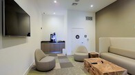 This single-floor commercial space offers a clean, modern design with a thoughtfully arranged lounge area that sets a professional yet comfortable tone. The lounge features contemporary pod-style seating chairs in neutral tones, paired with a rustic natural wood coffee table that adds an organic contrast to the otherwise sleek interior. A patterned carpet in muted greens and grays defines the seating area, providing a subtle visual anchor within the open layout.

A dedicated refreshment station lines one wall, outfitted with a dark countertop, warm wood cabinetry, and a professional-grade coffee machine, making it a convenient amenity for visitors and occupants alike. Recessed ceiling lighting illuminates the space evenly, complementing the crisp white walls and neutral color palette accented by warm taupe tones.

Restroom facilities are conveniently accessible directly from the lounge area, clearly marked with universally recognized signage. HVAC vents integrated into the ceiling and walls ensure climate control throughout. The overall ambiance is polished and inviting, reflecting a well-appointed professional environment suitable for a corporate office, medical facility, wellness center, or similar commercial use.