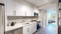 This efficiently designed single-floor unit offers a thoughtful layout encompassing a kitchen, living room, and bathroom. The modern kitchen features floor-to-ceiling white cabinetry, a herringbone-patterned tile backsplash, white quartz countertops, and a full suite of stainless steel appliances including a gas range, built-in microwave, and dishwasher. Light wood-tone flooring runs throughout, creating a cohesive and airy feel.

A corridor connects the kitchen to the living area, which is brightened by large floor-to-ceiling windows offering treetop views and abundant natural light. The living space is versatile, accommodating both a seating arrangement and a compact workspace, making it well-suited for modern living. Neutral walls provide a clean backdrop throughout.

The entryway includes a double-door closet offering practical storage, alongside a warm wood-toned entry door with a keypad lock. The bathroom is cleanly finished with a glass-enclosed walk-in shower lined in classic white subway tile, neutral stone-toned flooring, and chrome fixtures. The overall design aesthetic is contemporary and cohesive, with a neutral palette, clean lines, and modern finishes that maximize both function and comfort throughout the space.