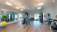 This fitness center offers a thoughtfully designed workout environment that balances functionality with an inviting aesthetic. The space features rich hardwood-style flooring throughout, providing a warm yet durable surface suited for a variety of exercise activities. Crown molding and soft sage-green walls lend an elevated, polished feel uncommon in typical gym settings.

Lighting is a standout element, with large drum pendant lights suspended from the ceiling complementing recessed lighting to ensure the space is consistently well-lit. Multiple windows along the walls allow natural light to pour in, creating an open and energizing atmosphere.

The workout floor is generously proportioned, offering ample room to move freely between equipment stations. Strength training is well-supported with a multi-station cable machine, a functional trainer, a weight bench, and a full rack of dumbbells neatly arranged along the wall. A floor-to-ceiling mirror runs along one side of the room, enhancing the sense of space and allowing users to monitor their form during exercise.

Ceiling fans are positioned throughout to promote airflow and comfort during workouts. The overall layout is clean and uncluttered, making navigation between equipment intuitive and efficient. This fitness center provides a comprehensive and comfortable setting for both strength and general training needs.