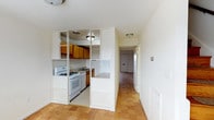 This two-floor space is defined by its consistent use of warm-toned parquet flooring, which runs throughout the rooms and lends a sense of cohesion and classic character to the interior. The layout is practical and well-organized, with a central corridor connecting the key areas of the home.

The kitchen is a functional and efficient space, partially divided from the adjoining area by a white open shelving unit that offers both storage and a degree of visual separation without fully closing off the space. Warm wood cabinetry lines the walls, complemented by a white gas range and oven, a sink, and a countertop pass-through that adds to the open feel. Flush-mounted ceiling lighting illuminates the area evenly.

The living and sleeping areas are generously proportioned, featuring clean white walls and the same parquet flooring that carries throughout. One room benefits from a large sliding glass door that allows an abundance of natural light to filter in, contributing to a bright and airy atmosphere. The rooms offer a blank canvas with neutral finishes, making them highly adaptable to a variety of uses and furnishing styles.

A hallway connects the rooms across both floors, providing a clear and navigable flow through the space. Overall, the interior presents a clean, understated aesthetic with practical features and a warm, light-filled ambiance.