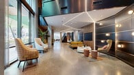 Located at 620 Merritt Ave in Nashville's vibrant Wedgewood-Houston neighborhood, this two-floor amenity facility offers a thoughtfully designed environment that blends modern aesthetics with functional versatility. Polished terrazzo flooring flows consistently throughout both levels, anchoring the space in a cohesive, upscale material palette.

The main floor greets visitors with a grand, open lobby featuring soaring ceilings, geometric linear LED lighting, and warm wood-paneled accents set against concrete and dark metal surfaces. Carefully arranged seating vignettes — including mustard-toned sofas, accent chairs, and circular coffee tables — create inviting social hubs. A striking marble reception desk with slatted wood backdrop serves as a refined focal point, while curated artwork and murals add creative energy throughout.

Floor-to-ceiling sheer-draped windows on the main level flood a lounge area with natural light and offer views of the surrounding Nashville streetscape. A dedicated maker studio features industrial-style worktables, adjustable stools, pegboard tool walls, and utility sinks — ideal for hands-on creative pursuits.

The second floor continues the design language with additional lounge seating areas, glass-partitioned spaces, pendant lighting clusters, and a reception counter with dark granite detailing. Connected by an open staircase, both floors together deliver a dynamic, amenity-rich environment suited for community engagement, collaboration, and relaxation.