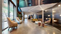 Located at 620 Merritt Ave in Nashville's vibrant Wedgewood-Houston neighborhood, this two-floor amenity facility offers a thoughtfully designed environment that blends modern aesthetics with functional versatility. Polished terrazzo flooring flows consistently throughout both levels, anchoring the space in a cohesive, upscale material palette.

The main floor greets visitors with a grand, open lobby featuring soaring ceilings, geometric linear LED lighting, and warm wood-paneled accents set against concrete and dark metal surfaces. Carefully arranged seating vignettes — including mustard-toned sofas, accent chairs, and circular coffee tables — create inviting social hubs. A striking marble reception desk with slatted wood backdrop serves as a refined focal point, while curated artwork and murals add creative energy throughout.

Floor-to-ceiling sheer-draped windows on the main level flood a lounge area with natural light and offer views of the surrounding Nashville streetscape. A dedicated maker studio features industrial-style worktables, adjustable stools, pegboard tool walls, and utility sinks — ideal for hands-on creative pursuits.

The second floor continues the design language with additional lounge seating areas, glass-partitioned spaces, pendant lighting clusters, and a reception counter with dark granite detailing. Connected by an open staircase, both floors together deliver a dynamic, amenity-rich environment suited for community engagement, collaboration, and relaxation.