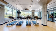 This generously sized fitness center offers a well-appointed, single-floor layout designed to accommodate a full range of workout activities. The space is defined by its industrial-modern aesthetic, featuring exposed concrete columns, visible ductwork along the ceiling, and warm hardwood flooring throughout that provides both visual warmth and a durable workout surface.

The cardio zone is a standout area, lined with rows of Star Trac treadmills and additional cardio machines positioned to face expansive floor-to-ceiling grid-pane windows. These large windows flood the space with natural light and frame views of lush outdoor greenery and an exterior terrace, creating an energizing atmosphere for exercise. Wall-mounted flat-screen televisions are positioned throughout for additional entertainment.

A dedicated cycling area accommodates Stages spin bikes alongside a wall-mounted screen and direct access to natural light from oversized windows. An open studio space provides a clear, flexible area ideal for stretching, group classes, or functional training, with mirrored walls offering views into the adjacent cardio section. The entry area features polished concrete flooring, blue accent walls, and a welcoming transition into the main fitness floor, completing a cohesive and functional facility.