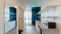 This thoughtfully designed single-floor unit combines modern aesthetics with practical, accessible living. The kitchenette is a standout feature, showcasing bold teal accent walls that contrast sharply with crisp white cabinetry, open bracket shelving, and granite-topped counters. Equipped with a stainless steel compact refrigerator, microwave, and a black-fixtures sink, the kitchenette also includes a built-in desk area with a window overlooking the surrounding neighborhood — ideal for daily tasks. An adjacent pantry or storage area with open shelving adds generous organizational space. The sleeping area is clean and minimalist, featuring a platform bed with built-in drawer storage beneath and a wall-mounted mini-split HVAC unit for individual climate control. Neutral tones and recessed lighting keep the room bright and functional. The bathroom is fully ADA-compliant, outfitted with a roll-in shower, handheld showerhead, multiple grab bars in both chrome and matte black finishes, and dark tile flooring. A separate living or flex space benefits from large windows fitted with horizontal blinds that invite ample natural light. Throughout the unit, consistent light flooring, clean white walls, and modern black hardware create a cohesive, contemporary feel.