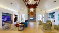 Located at 2015 Gum Branch Rd in Jacksonville, NC, this thoughtfully designed single-floor community clubhouse spans an impressive footprint, offering residents a diverse array of social and functional spaces. The grand living room immediately commands attention with its soaring vaulted ceiling, rich wood beam accents, a floor-to-ceiling stacked stone fireplace, and a striking starburst chandelier. Arched windows flood the space with natural light while providing views of the surrounding community. Wide-plank light hardwood flooring flows throughout, contributing to a warm, welcoming ambiance. The dining area is a standout, featuring an eye-catching floor-to-ceiling swirling blue-and-white marbled mural wall, a teal ceiling, and bistro-style tables with woven rattan chairs. The adjacent kitchen is finished with white cabinetry, stone countertops, and a herringbone tile backsplash. The game room offers a full-size billiards table beneath modern pendant lighting with a wet bar and dark cabinetry nearby. Interconnecting hallways showcase patterned tile flooring, frosted glass French doors, and a dedicated media room with geometric wallpaper. A practical mailroom and package locker area, complete with vending machines and a skylight, rounds out this well-appointed community facility.