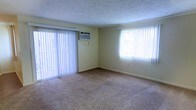 This single-floor space presents a straightforward and functional layout designed for comfortable everyday living. The main room is a standout feature, offering a generous open area finished with wall-to-wall carpeting in a neutral beige tone that adds warmth underfoot. Two large windows dressed with white vertical blinds allow an abundance of natural light to fill the room, creating a bright and airy atmosphere. A wall-mounted air conditioning unit is conveniently installed beneath one of the windows, ensuring climate control throughout the warmer months. The walls are painted in a soft, light yellow hue that complements the neutral flooring and contributes to the room's overall sense of calm and openness. White baseboards frame the perimeter of the room, adding a clean, finished look. The textured ceiling adds a subtle visual detail to the space. The absence of interior clutter allows the room's proportions to be fully appreciated, making it well-suited for a variety of furniture arrangements and uses. Overall, this space offers a clean, practical environment with ample natural light and essential climate features.