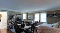This single-floor space offers an open, combined living and dining layout that maximizes functionality within a compact footprint. The room features warm, neutral-toned walls complemented by classic crown molding, lending a touch of traditional character to the interior.

The dining area is anchored by a dark wood table with matching X-back chairs, paired with a large wooden bowl centerpiece, creating a practical yet inviting spot for meals and gatherings. Adjacent to this, the living area is furnished with a sofa and additional seating, making it a comfortable space for relaxing.

An entertainment setup with a flat-screen television and media console is positioned along one wall, while a white writing desk provides a dedicated workspace nearby. A cork board above the desk adds a personal, organized touch to the study nook. Decorative banners and personal touches throughout the space reflect a lived-in, homey character.

Multiple windows along the walls allow generous natural light to fill the room, creating a bright and airy atmosphere throughout the day. A small potted plant near the windows adds a refreshing natural element. Carpeted flooring throughout contributes to the overall warmth and comfort of this well-utilized, single-level living space.