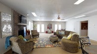 This single-floor space is centered around a spacious and welcoming living area designed for comfort and functionality. The room is illuminated by multiple skylights embedded in a textured ceiling, complemented by recessed lighting and a ceiling fan that together create a bright, airy atmosphere throughout the day.

A classic brick fireplace anchors one wall, adding warmth and a traditional focal point to the space. Flanking it are tall decorative vases, enhancing the room's character. On the opposite side, a wall-mounted flat-screen television provides a modern entertainment element.

The living area is furnished with plush, oversized sofas and accent chairs arranged around a decorative area rug with a bold floral pattern, creating a cozy and inviting gathering space. A rounded ottoman serves as a central coffee table, adding a relaxed, casual feel.

Multiple sets of French doors line the walls, flooding the interior with natural light while offering direct access to adjacent areas or the outdoors. Light-toned flooring runs throughout, contributing to the open and expansive feel of the space. The overall ambiance is warm and livable, blending traditional design elements with comfortable, functional furnishings.