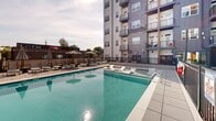 Located at 1829 N Meridian St in Indianapolis, Indiana, this outdoor amenity courtyard is the centerpiece of a contemporary multi-story residential complex. The space is designed to maximize outdoor living, combining a resort-style swimming pool with a spacious surrounding sun deck finished in large-format pavers. The pool area is outfitted with rows of lounge chairs and striped umbrellas, providing ample space for relaxation. Integrated in-pool sun shelf loungers add a resort-caliber touch to the aquatic experience. Encircling the pool, wrought-iron fencing with clean black metalwork defines the space while maintaining sightlines throughout the courtyard. A dedicated green lawn area with seating provides a lush alternative to the hardscaped pool deck, complemented by a shaded pergola structure ideal for outdoor dining or gathering. Raised planters filled with ornamental grasses and sculpted evergreens add structured greenery throughout. The courtyard is framed on all sides by the multi-story building façade, which blends gray siding, exposed brick, warm wood accent panels, and private balconies. String lights and pathway lanterns enhance the ambiance for evening use. Entry to the courtyard is provided through glass double doors with a prominent warm-toned wood surround, offering a welcoming transition between interior and exterior spaces.