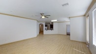 This single-floor space features an expansive open-plan living area that immediately draws attention with its light wood-style flooring and crisp white walls accented by tan baseboard trim and crown molding. The main living area is anchored by a ceiling fan with a built-in light fixture, contributing to air circulation and ambient lighting alongside a recessed fluorescent light fixture visible toward the back of the space. A ceiling-mounted HVAC vent ensures year-round climate comfort.

A defining architectural feature is the arched doorway that connects the living area to a hallway, adding a classic design touch. The kitchen is partially open to the living space via a raised breakfast bar counter, offering a convenient pass-through layout. Dark cabinetry and a stainless steel refrigerator are visible in the kitchen area, providing a contrast to the otherwise neutral palette throughout.

Additional doorways are visible on either side of the space, suggesting connectivity to further rooms. The overall ambiance is clean, neutral, and versatile, making it well-suited for a variety of furniture arrangements and interior styles.