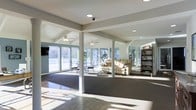 This single-floor space offers an open, airy layout filled with abundant natural light streaming through large floor-to-ceiling windows that line the exterior walls. The interior is defined by clean architectural details, including white support columns, vaulted ceiling sections with exposed beams, and recessed lighting complemented by multiple ceiling fans throughout. The space is organized into several functional zones that flow seamlessly into one another. A sunlit seating area near the windows features casual dining furniture, ideal for relaxed gatherings. The central lounge area is furnished with comfortable reclining chairs and arranged around an open carpeted section, providing a welcoming atmosphere. Toward the back of the space, fitness equipment is visible, indicating a dedicated exercise area. A dark wood shelving unit adds a warm, decorative accent along one wall. The flooring transitions from polished tile in the main entry zone to carpet in the lounge area, subtly delineating each section. The overall ambiance is light, clean, and versatile, making this space well-suited for a variety of community or recreational purposes.