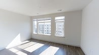 This single-floor space showcases a clean, contemporary design characterized by a minimalist aesthetic and an open layout. The main living area is finished with warm-toned hardwood flooring that contrasts elegantly against the crisp white walls, creating a bright and inviting atmosphere. Three large windows dominate one wall, allowing abundant natural light to stream in throughout the day, casting dramatic sunlit patterns across the floor. The windows offer direct views of the surrounding urban environment, connecting the interior to the cityscape outside. Ceiling-mounted HVAC vents are integrated discreetly into the white ceiling, contributing to the space's clean, uncluttered appearance. The neutral color palette and smooth wall finishes provide a versatile backdrop suitable for a wide range of interior styles. White baseboards frame the perimeter of the room, adding a refined finishing detail that complements the overall modern sensibility. The space presents a well-lit, move-in ready environment with a straightforward, functional layout ideal for flexible use.