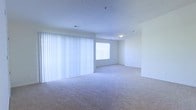 This single-floor space presents a clean, open layout finished in a neutral palette that lends a bright and airy atmosphere throughout. The main living area is generously proportioned, featuring wall-to-wall carpet in a light beige tone that adds warmth underfoot. A large set of vertical blinds spans one full wall, diffusing natural light softly across the room while offering privacy and light control. An additional window in the adjoining area further enhances the natural brightness of the space. The white walls and white ceiling contribute to the overall sense of openness, reflecting light evenly throughout. Ceiling-mounted lighting provides supplemental illumination. The open floor plan allows for flexible furniture arrangement and a seamless flow between areas. The space is vacant and move-in ready, offering a blank canvas for personalization. Overall, the unit maintains a simple, functional design with an emphasis on light, space, and ease of use.