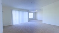 This single-floor space presents a clean, open layout finished in a neutral palette that lends a bright and airy atmosphere throughout. The main living area is generously proportioned, featuring wall-to-wall carpet in a light beige tone that adds warmth underfoot. A large set of vertical blinds spans one full wall, diffusing natural light softly across the room while offering privacy and light control. An additional window in the adjoining area further enhances the natural brightness of the space. The white walls and white ceiling contribute to the overall sense of openness, reflecting light evenly throughout. Ceiling-mounted lighting provides supplemental illumination. The open floor plan allows for flexible furniture arrangement and a seamless flow between areas. The space is vacant and move-in ready, offering a blank canvas for personalization. Overall, the unit maintains a simple, functional design with an emphasis on light, space, and ease of use.
